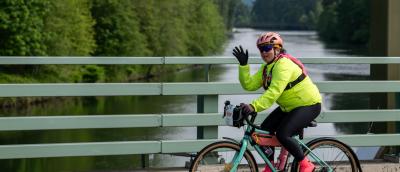 Woman on a bike crossing a bridge over a river waves at the camera.